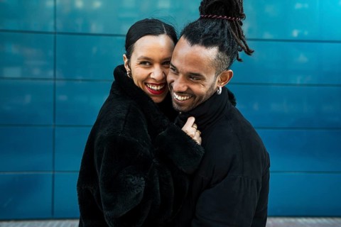 A multicultural couple stands close in a loving embrace, smiling warmly. The urban background adds