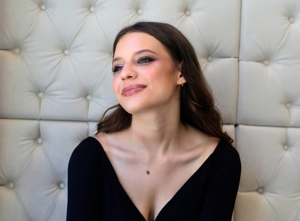 A woman with vibrant makeup and a subtle smile gazes thoughtfully. She is in an elegant setting