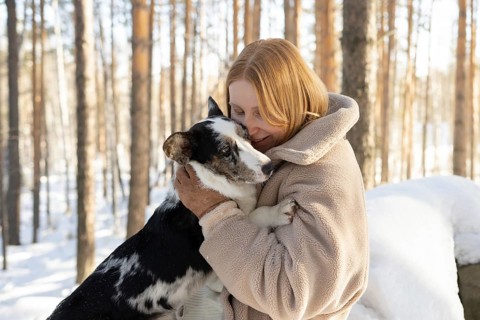 A redhead woman warmly embraces her dog, possibly a Border Collie mix, in a snowy forest setting.