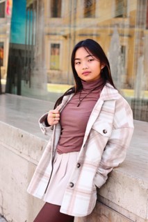 Young woman with a stylish plaid coat and pink blouse leaning against a stone ledge in an urban