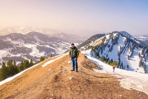 A hiker stands on a snow-covered mountain ridge with extensive mountain views under clear skies,