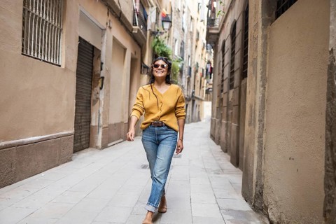 Latin woman in casual attire joyfully walks down a narrow, sunlit city street lined with old