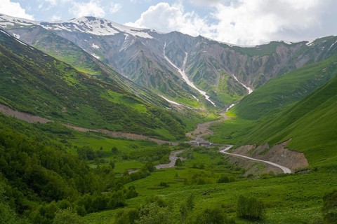 Green valley with snow-covered mountains under cloudy sky and a winding road, near Zagari Pass,