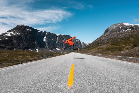A traveler in a red jacket leaps mid-air on an open mountain road in Norway Framed by dramatic