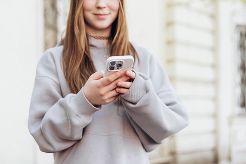 Teenage girl holding a smartphone while outdoors, dressed in a casual gray sweatshirt. The focus is