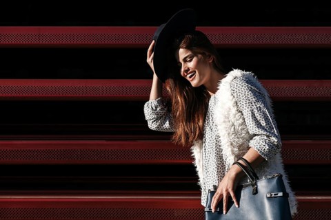 A fashionable woman poses outdoors on a sunny day, wearing a black hat, patterned shirt, and fur