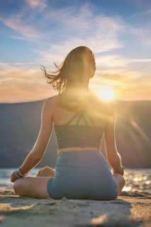 A woman sits in Padmasana on the beach, meditating with Chin mudra as the sun sets, creating a