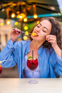 Vertical photo of a caucasian woman enjoying biting the citrus garnishing a cocktail in an outdoor