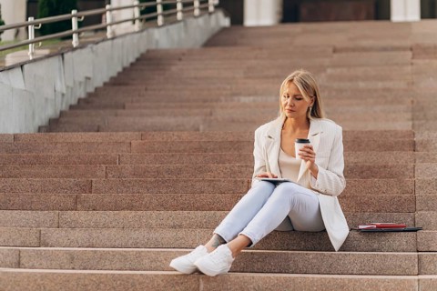 A businesswoman in casual business attire sits on outdoor steps, holding a coffee cup and using a