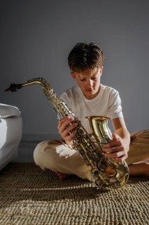 A young musician sits indoors in soft natural light, examining a saxophone. The scene captures
