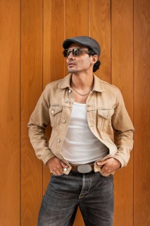 A fashionable man wearing sunglasses, a tan jacket, and a cap stands confidently against a wooden