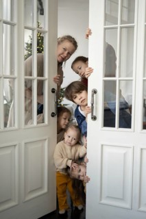 A cheerful family of five peeks around a white glass door, revealing bright smiles and curious