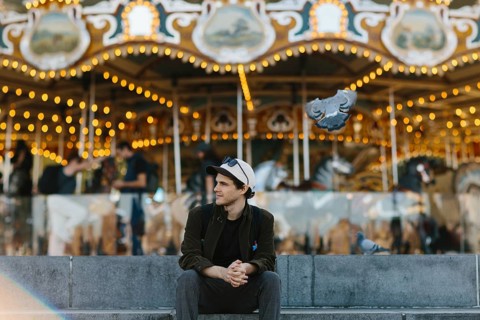 A young man sits casually in front of a brightly lit carousel in Brooklyn Bridge Park. Pigeons fly