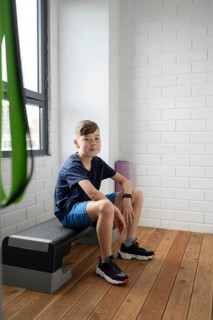 A young boy sits on a bench in a modern, brightly lit room with wooden floors and white brick