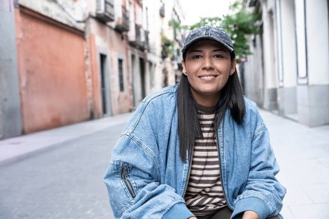 Latina woman wearing a striped shirt and denim jacket smiles confidently while sitting on a vibrant