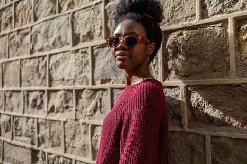 A stylish woman in a burgundy sweater wearing sunglasses stands confidently against a textured