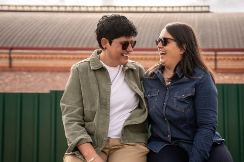 A joyful curvy lesbian couple shares a light heart moment outdoors, sitting closely and wearing