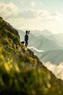 A man takes a break from trail running to hydrate, standing on a scenic mountain path with distant