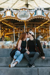 A couple shares a kiss on stone steps in front of a brightly lit carousel in Brooklyn Bridge Park.
