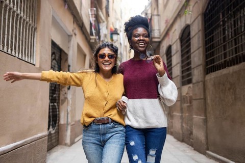 Two friends joyfully walk through a narrow street, one wearing sunglasses and the other holding