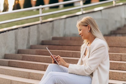 A businesswoman in casual business attire sits on outdoor steps while using a tablet. The setting