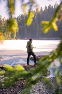 Man standing by a frozen lake in a wintery forest, framed by green branches, Glaswaldsee, Bad