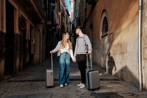 A young couple walks hand in hand down a narrow street, luggage in tow, as they search for their