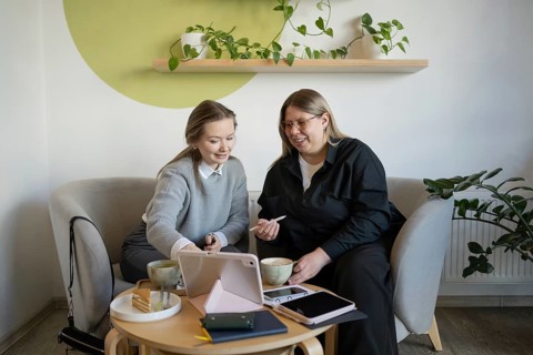 Two women sit on cozy chairs in a cafe, sharing conversation over coffee. A tablet rests on the