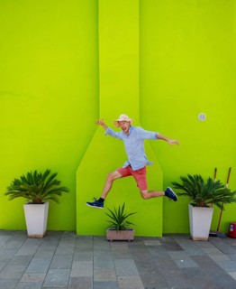 Young man jumping over plant pot in front of green house wall, green house facade with flowers,