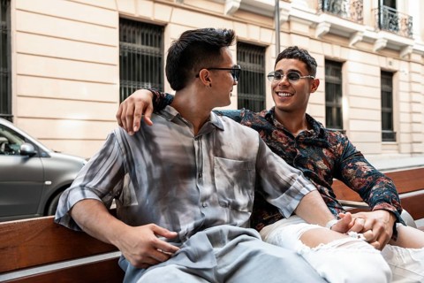 A cheerful latin gay couple sits close together on a bench, laughing and enjoying a sunny day in