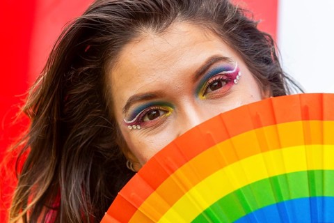 A woman with vibrant rainbow makeup partially conceals her face with a pride-themed fan. Her