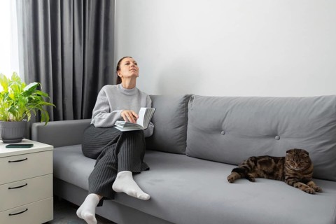 A woman sits on a gray sofa, holding a book, looking peaceful and contemplative. Beside her, a cat