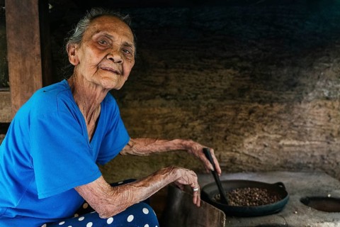 An elderly woman in a blue shirt sits by a rustic stove, roasting balinese coffee beans in a pan.