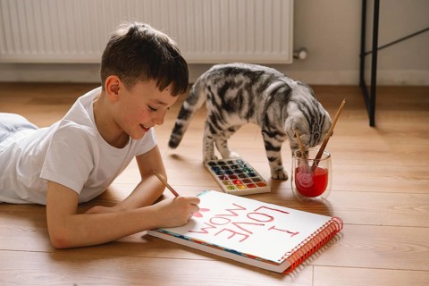 A boy lies on the floor, drawing a Mother's Day card with vibrant paints. His dedicated expression