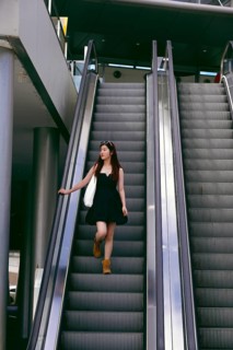A young asian woman in a black dress and boots descends an escalator in an urban setting. She