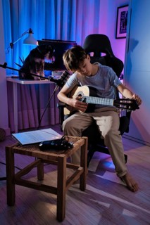 A young boy tuning his guitar in a cozy, blue lit room, with a microphone and sheet music nearby. A