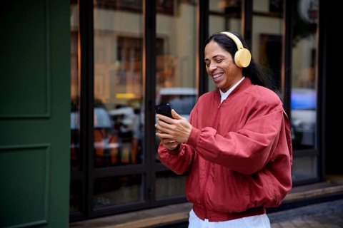 A man in a red jacket, wearing yellow headphones, smiles while looking at a smartphone. Captured