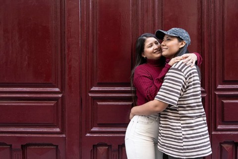 A Latin lesbian couple shares a loving embrace against a deep red wooden backdrop. They exude