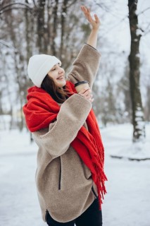 A cheerful young woman smiles as she lifts her hand in a snowy park, dressed warmly with a red