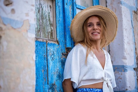 A smiling woman in a straw hat leans against a rustic blue door in Trypiti, Milos Island, Greece.