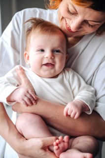 Loving mother cradles smiling newborn baby girl at home, both wearing white clothes. The baby's