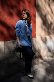 A stylish woman in a blue shirt leans against a red and beige wall, gazing into the distance. The