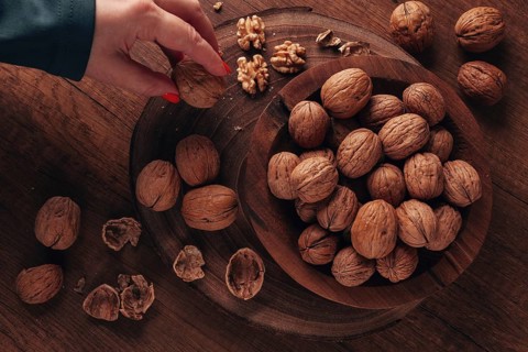 A woman's hand taking a walnut from a wooden bowl filled with walnuts. There are shells and walnuts