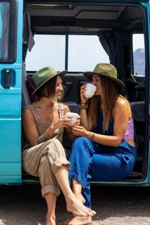 Two lesbian women enjoy leisure time by a camper van, sipping coffee and wearing casual attire.