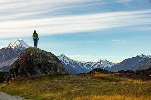 A woman stands on a rocky outcrop overlooking Mount Cook in New Zealand, surrounded by stunning