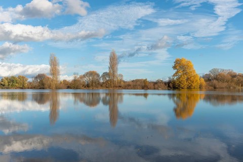 The river flows flat and reflects clouds and mountains under a clear sky. Autumn trees draw the
