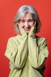 A joyful Asian Chinese senior woman with gray hair and a yellow sweater poses cheerfully against a