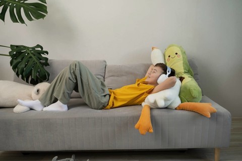 A young boy in a yellow shirt lies comfortably on a grey sofa, wearing headphones and surrounded by