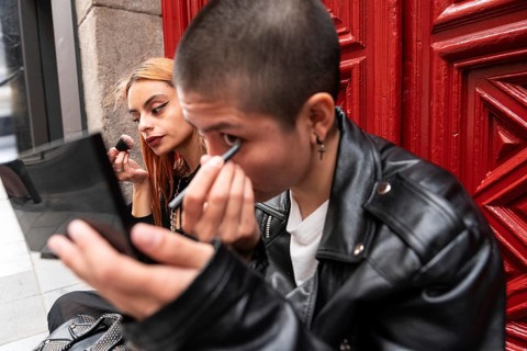 A lesbian couple sits together in front of a red door, each applying makeup with focus and care,