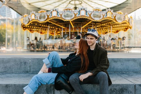 A young couple sits on steps in front of a brightly lit carousel in Brooklyn Bridge Park. They